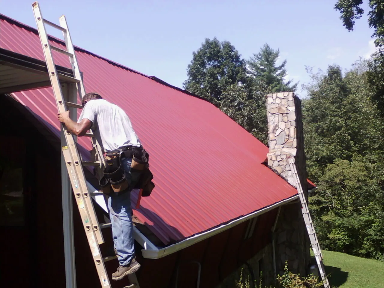 Roofer on a ladder beside a red metal roof and stone chimney — Soffit & Fascia Repair by Roofing Brothers in Pottersville
